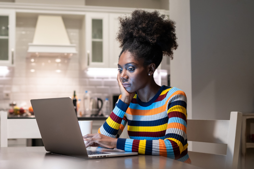Black woman, wearing a multicolored striped sweater, using laptop at home while sitting at the kitchen table.